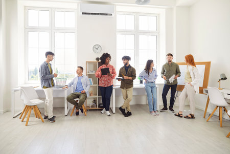 Full length photo of a group of young diverse business people men and women chatting after meeting. Company employees or group of staff talking in modern office during a break discussing work projectの写真素材