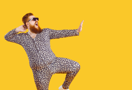 Funny eccentric man dressed in leopard print pajamas dancing and fooling around on orange background. Cheerful bearded man in sunglasses having fun at copy space. Web banner.の写真素材