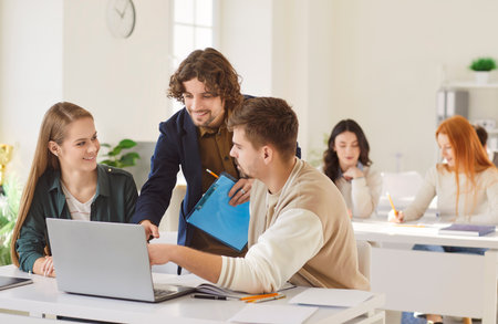 Young business friends, coworkers, students using laptop online at work table, talking discussing project ideas for learning strategy, planning startup, studying together, teacher helping to explainの写真素材