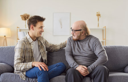 Happy elderly father enjoys leisure and conversation with adult son on a sofa at home, focusing on family love and fathers day bonding in a cozy setting with a sense of togetherness and affection.の写真素材