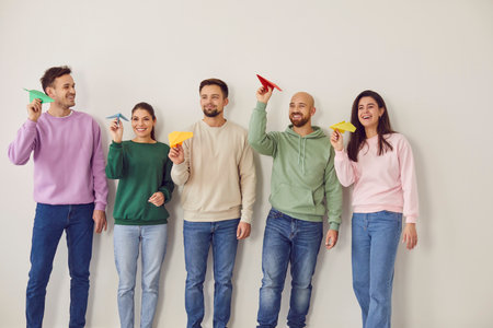 Team of happy smiling people flying paper planes together. Cheerful male and female friends in casual sweatshirts and jeans standing by white wall holding colorful airplanes. Dreaming, success conceptの写真素材