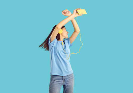 Happy millennial girl dance isolated on blue studio background listen to music in earphones using...