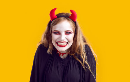 Young scary Caucasian woman with white make-up and devil horns showing teeth invites you to Halloween party celebrations on night of October 31 standing in yellow studio. Hallowmas, All HallowDayの写真素材