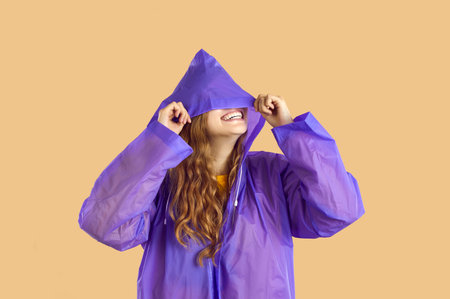 Smiling woman with blondy wave hair in purple raincoat and hood on her eyes is posing on yellow background, happy halloween concept. She is looking up through the transparent hood on rainy sky.の写真素材