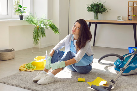 Portrait of a young woman sitting on the floor in the living room at home next to vacuum cleaner tired of doing housework. Exhausted girl vacuuming carpet in house frustrated with much houseworkの写真素材