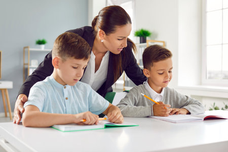 Young professional Caucasian woman teacher stands behind boys sitting at school desk, helps children complete task for pupils of lower grades located in bright classroom. Extracurricular educationの写真素材