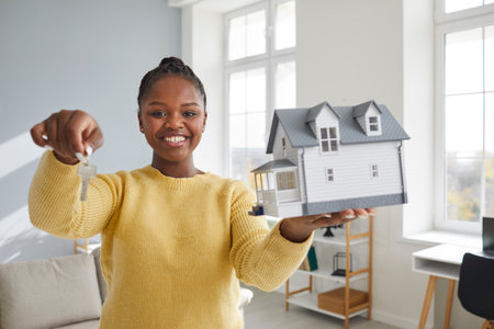 Portrait of a happy smiling woman at her new home. Cheerful joyful beautiful young African American woman holding a house model and showing the keys to her new property. Real estate conceptの写真素材