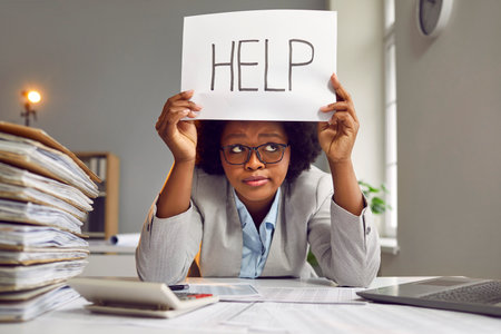 Tired, busy, sad African American woman in a suit and glasses sitting at an office desk with a load of paperwork and holding a paper sign with the word HELP. Working in the office, workload conceptの写真素材