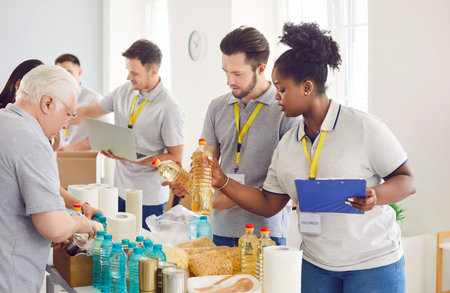 Diverse volunteers group sorting donations of foodstuffs in cardboard boxes for charity food drive. Mature and young people volunteering in food bank working in charitable foundation together.の写真素材