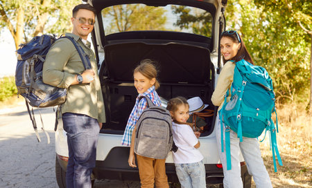 Happy family with backpacks by car trunk preparing for road trip. Parents and children pack luggage for outdoor travel and vacation on a sunny road near countryside. Concept family adventure.の写真素材