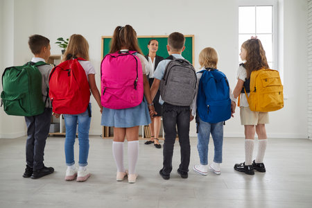 Classmates standing back around the friendly teacher at the classroom before lesson or on first school day and listening what she talking about. Education, primary school and back to school concept.の写真素材