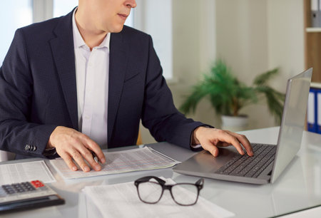 Cropped shot of businessman working on financial documents and using laptop in office. Male...