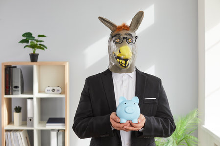 Portrait of a funny man in a suit wearing animal donkey mask on head standing in the office holding piggy bank in hands. Savings, investment and business profit, entertainment and fun concept.の写真素材