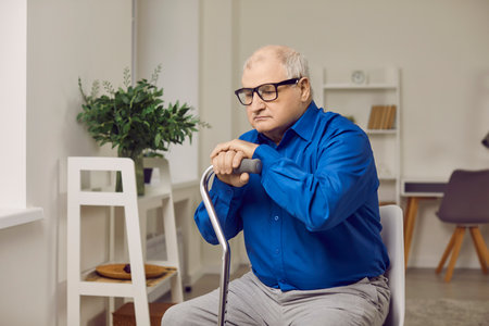 Portrait of a senior man sitting at home in rehabilitation with his crutch and looking sad in eyeglasses. Gray-haired elderly man with walking stick sitting on a chair in the living room.の写真素材