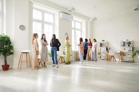 Group of diverse women standing in small groups by windows in bright spacious office, talking to each other during meeting. Multinational team of female coworkers or friends discussing work issues.の写真素材
