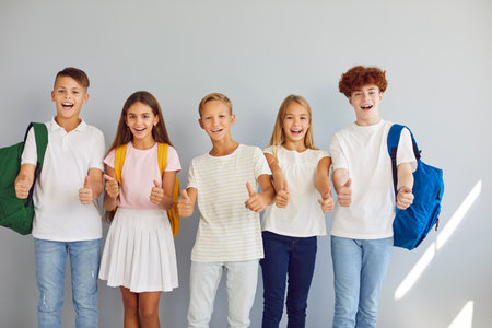 Happy elementary school kids give thumbs up. Group portrait of joyful smiling preteen male and female children in casual wear with backpacks standing by grey studio wall and showing thumbs up symbolsの写真素材