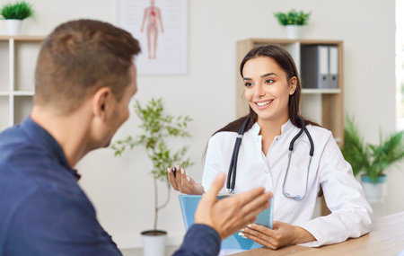 Friendly female physician or cardiologist sitting at desk in office, listening to patient...