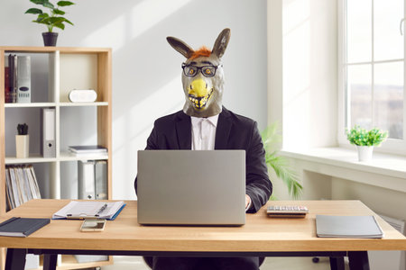 Funny business man and company employee wearing suit and animal donkey mask sitting at the desk on his workplace and working on a laptop in office. Entertainment, fun and fools day concept.の写真素材