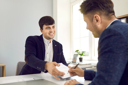 Positive smiling manager giving contract to sign to his client on business meeting in office sitting at desk. Business transaction, conclusion of contract, provision of services, office work concept.の写真素材