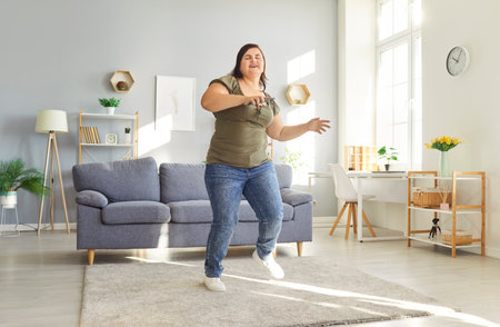Happy overweight young woman dancing joyfully in her living room, enjoying physical activity and positive energy. Smiling female listening to music, enjoying free time and having fun at home.の写真素材