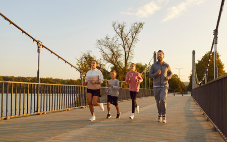 Family running in park, children jog together. Parents set an outdoor fitness pace on a bridge at...