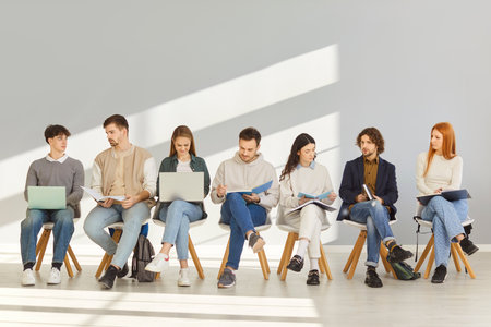 Group of young people sitting in row on chairs in bright room, focused on reading, writing and using laptops. Students or job seekers engaged in study or work activities, preparing for job interview.の写真素材