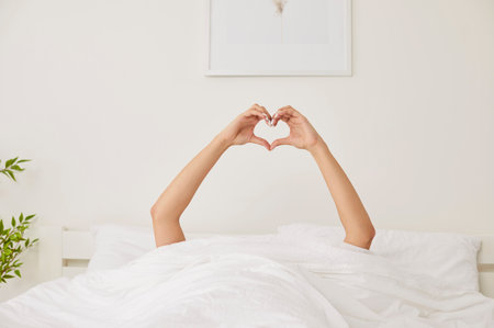 Human hands showing heart shape symbol from under blanket. Happy young woman lying in bed waking up early in morning in sunny bedroom, demonstrating love and gratitude to new day. Good morning conceptの写真素材