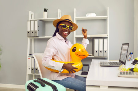 Happy smiling african american woman sitting at the desk on workplace at office in a beach rubber ring with suitcase and booking tickets for summer vacation online via laptop.の写真素材
