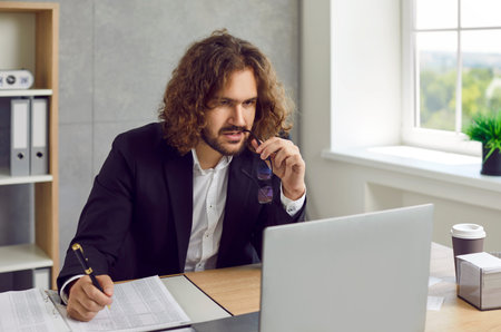 Portrait of young attractive business man with long hair working on a laptop at office or at home,...