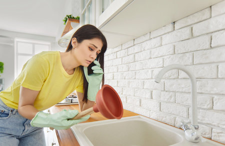 Young girl talking by the phone advising how to unclog a sink drain. Girl trying to clean sink using plunger standing in the kitchen at home. Housekeeping and household work concept.の写真素材