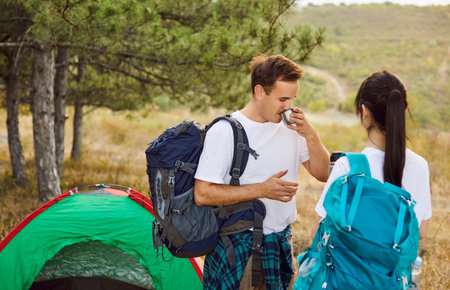 Couple of tourists camping with backpacks near forest camp tent. Man drinking hot drink from mug while woman holding thermos, people relaxing after hike in pine woods and weekend hiking travelの写真素材