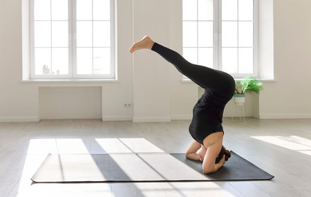 Yoga headstand inversion on mat demands balance. A woman practices a controlled variation by bright windows, engaging core. Light highlights alignment. Strength and focus define the concept.の写真素材