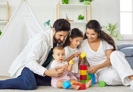 Happy family, parents and 2 children, playing with colorful toy blocks indoors on carpet in living room. Young mother, father, older kid and baby together play with plastic building blocks at homeの写真素材