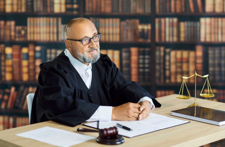 Judge sits thoughtfully with gavel and scales. Wearing robe, the magistrate listens at a desk among law books, weighing trial pleas and drafting a verdict. Justice, legal law, and authority concept.の写真素材