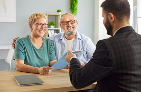 Smiling senior couple consults a financial advisor in a bright office, reviewing paperwork and discussing contract about investments and insurance going to sign documents during a meeting.の写真素材