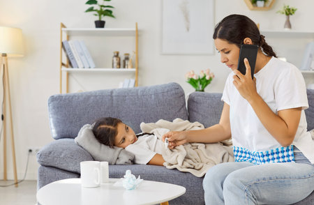 Sick child with fever watching mother. Caring parent on couch shows thermometer and calls doctor about virus by smartphone at home. Girl lies under blanket. Parental care during childhood illness.の写真素材