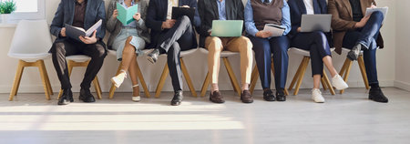 Candidates waiting interview with resumes for hiring. People on chairs using laptops in a bright office lobby awaiting team review and job tests. Symbol of recruitment and hiring process.の写真素材
