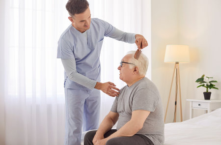 Young caregiver assisting senior man with his daily grooming routine in bright bedroom. Friendly male medical nurse helping elderly patient sitting on bed in nursing home to brush his hair in morning.の写真素材