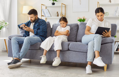 Child girl and parents on couch at home. Parents on smartphone and tablet as daughter sits upset and...