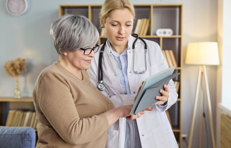 Doctor with senior patient using tablet. At home during visit they review test results and explain next steps, delivering advice support and care. Clear concept of helpful home consultation.の写真素材