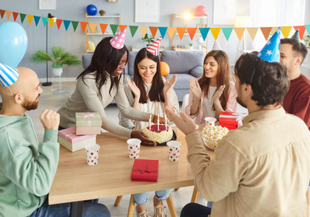 Group of cheerful young people wearing party hats congratulating birthday woman with cake. Happy...