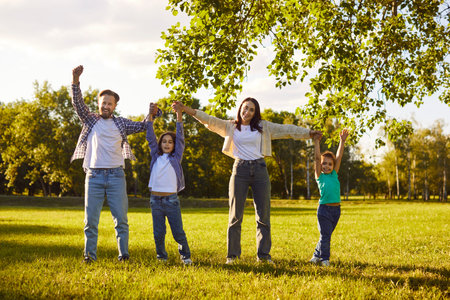 Happy family having fun standing in summer park together enjoying sunny day in nature. Mother, father and two cheerful children holding hands walking on green grass outdoors at sunset.の写真素材