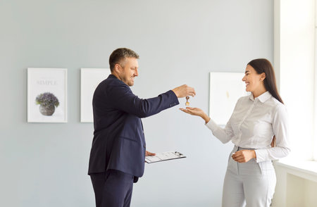 Positive male real estate agent standing in bright room, handing keys from new house to happy...