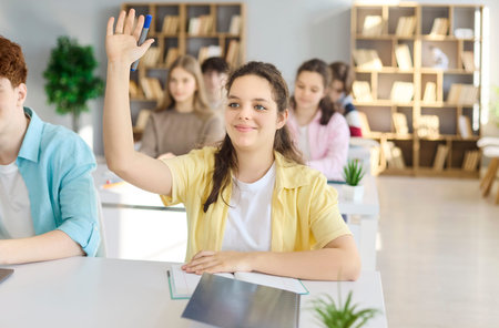 Student girl in classroom raise hand with question. Engaged teen listens with peers at school desks...