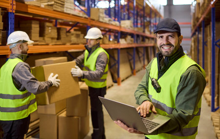 Warehouse inventory laptop worker boxes shelving. Supervisor updates stock counts as team moves...