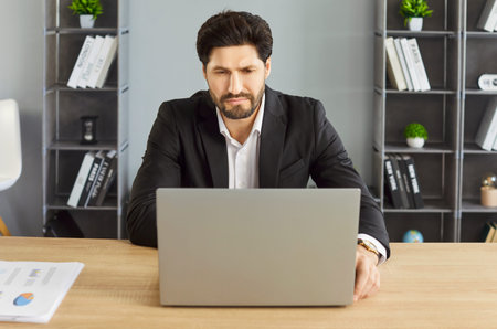 Serious businessman in formal suit sitting at office desk and working on laptop. Focused male...