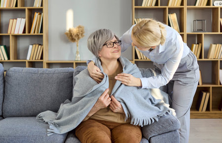 Senior woman with caregiver sharing home support. A warm blanket around her shoulders as they smile,...
