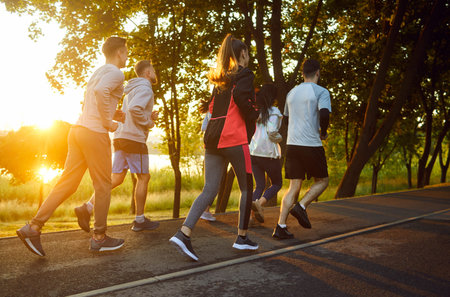 Full-length view, group of young people running along scenic route in summer park during sunset. Men...