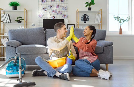 Couple cleaning at home with vacuum and gloves. Smiling partners high five on the floor by the sofa in aprons, with bucket, spray and vacuum visible, celebrating. Teamwork and clean living theme.