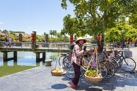 Hoi An, Vietnam 2024, August 12: A woman in traditional attire carries fruit-filled baskets along the riverside in Hoi An, Vietnam, with the iconic bridge and colorful buildings in the backgroundの写真素材